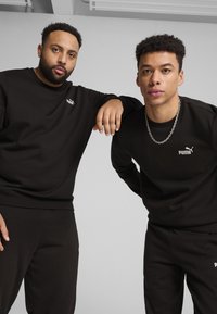 Felpe e pantaloni della tuta neri realizzati in materiale morbido, con loghi bianchi di Puma sul petto. Entrambi gli outfit presentano una vestibilità ampia.