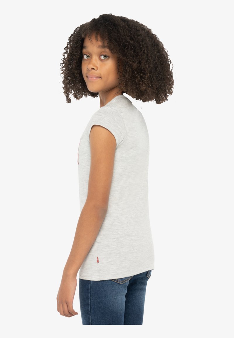 T-shirt gris à manches courtes en matériau doux, avec un petit logo sur la poitrine et une étiquette rouge près de l'ourlet.