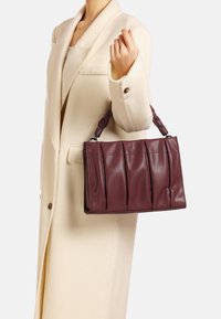 Borsa a mano in pelle malva con design plissettato, struttura solida e manico intrecciato, tenuta da una persona in un cappotto color crema.