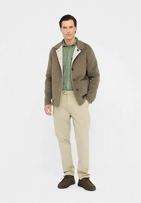 Giacca in misto lana marrone con colletto crema, camicia verde con bottoni, pantaloni beige e scarpe marroni, che presenta un look casual stratificato.