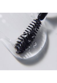 Envase de silicona transparente que contiene una varita de rímel negra, con cerdas y gotas de agua. Superficie lisa con forma curva.