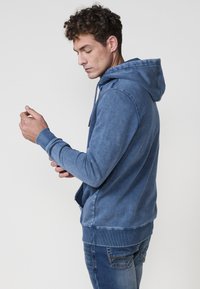 Modrá mikina vyrobená z jemnej látky, s kapucňou na šnúrku, rebrovanými manžetami a spodným okrajom. Viditeľné sú bočné vrecká. Nosí sa s modrými džínami.