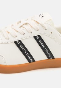 Bílé kožené tenisky s krémovými tkaničkami, černými bočními pruhy s nápisem "Armani Exchange" a světlou hnědou gumovou podrážkou.