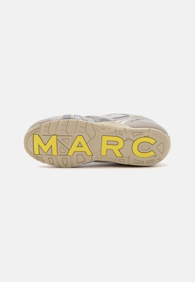 Baskets beiges vues de dessous, avec de grandes lettres jaunes formant "MARC" sur la semelle en caoutchouc texturée, sur fond blanc.