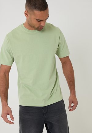 Mężczyzna w jasnozielonym t-shircie z krótkim rękawem i ciemnych dżinsach, patrzący w dół, stoi na tle jednolitego jasnego tła.