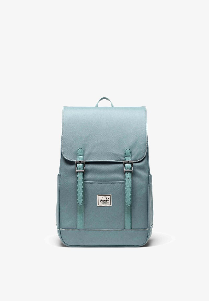 Una mochila de tela color verde azulado presenta un cierre con solapa con dos correas ajustables, un bolsillo delantero y un parche con la marca. Textura suave con detalles cosidos.