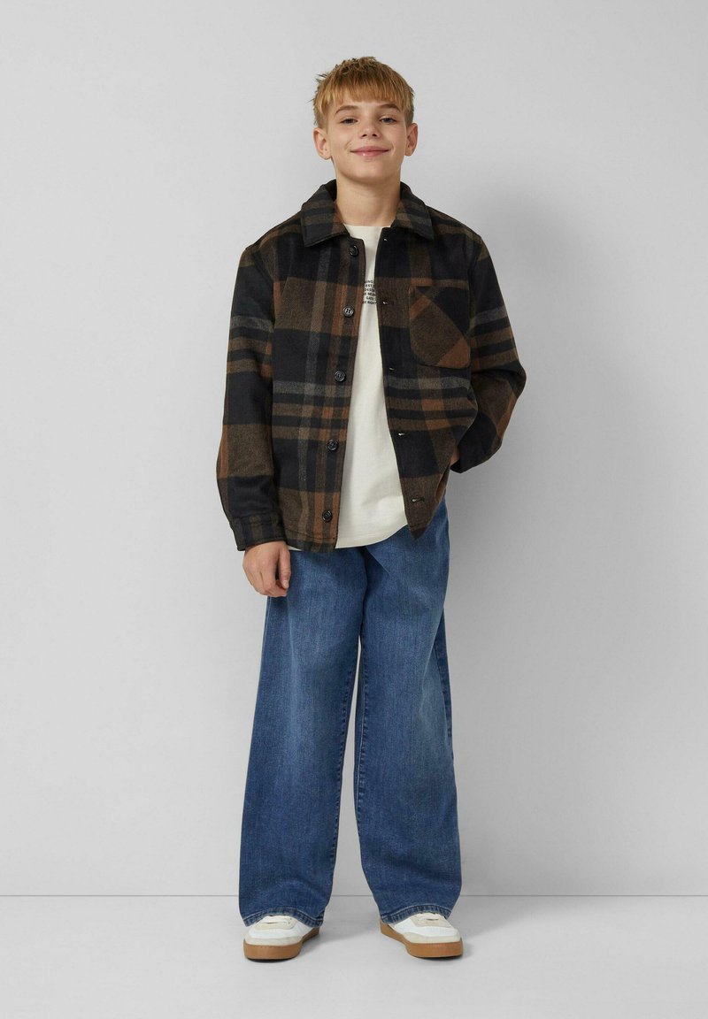 Smiling boy stands against gray wall wearing a black and brown checkered jacket, white shirt, wide blue jeans, and white sneakers.