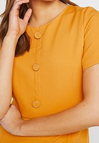 Vestido de manga corta en tejido amarillo brillante, con botones circulares dorados en la parte frontal, con una textura suave y un corte recto.