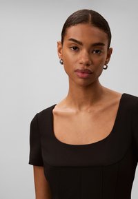 Jeune femme aux cheveux lisses tirés en arrière, portant des boucles d'oreilles argentées en anneaux et un top noir à col carré, sur un fond gris clair uni.