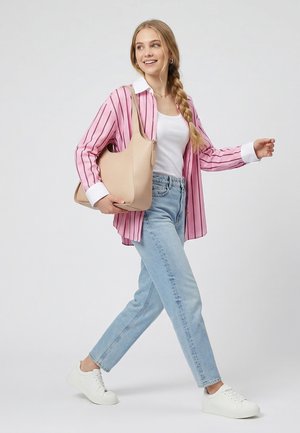 Mladá žena s dlouhým copy chodí oblečená v růžovém pruhovaném tričku, bílém topu, světle modrých džínách, bílých teniskách a nese béžovou kabelku přes rameno.