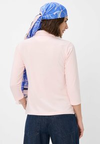 Lys pink langærmet skjorte med høj halsudskæring, der har en glat tekstur og sideslidser. Blå mønstret tørklæde bundet bagpå.