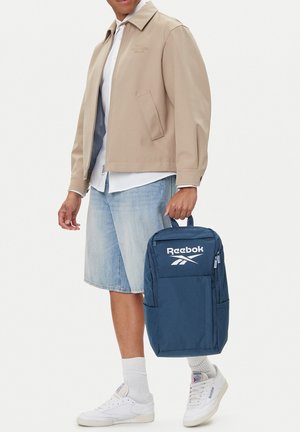 Persona con chaqueta beige, camisa blanca, pantalones cortos de mezclilla, zapatillas blancas, sosteniendo una mochila Reebok azul marino por la asa superior.