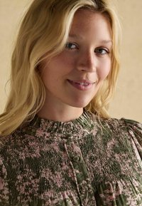 Noor blondidega noor naine kannab kõrge kaelusega lillekujulist pluusi roheline ja roosa tooni, naeratab õrnalt beežil taustal.