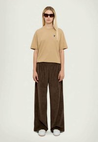 Beige cotton t-shirt with small navy logo, paired with loose, dark brown velour pants and white sneakers. Stylish sunglasses complete the look.