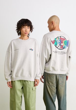 Gray oversized sweatshirt with multicolor "Vacation Club" graphic on the back. Model paired with green cargo pants. Casual fabric texture.