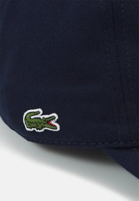 Boné de beisebol azul-marinho com tecido texturizado, apresentando um pequeno logotipo de crocodilo verde bordado e contornado a branco.