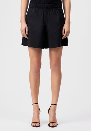 Kobieta ubrana w czarne szorty z wysokim stanem i elastycznym paskiem oraz czarne sandały na obcasie z paskiem wokół kostki, stojąca na jednolitym tle.