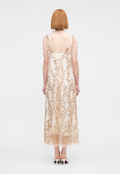 Μακρύ, αμάνικο φόρεμα με διαφανές επικάλυψη, με λεπτομερή χρυσή κεντητική φύλλων σε ανοιχτό φόντο. Απαλό, ρέον ύφασμα, μήκους έως τους αστραγάλους.