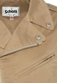 Giacca di suede beige con tasca zip in argento, bottoni a pressione in metallo incisi "Schott" e etichetta in tessuto bianco che mostra il marchio e la taglia piccola.