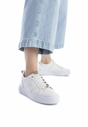 Zapatilla blanca con un diseño de malla texturizada, con detalles en hardware dorado y una suela de goma gruesa. Combinada con unos pantalones vaqueros de pierna ancha en azul claro.