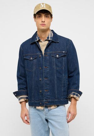 Young man wearing a beige cap, beige sweater, blue plaid shirt, dark blue denim jacket, and light blue jeans standing against plain background.