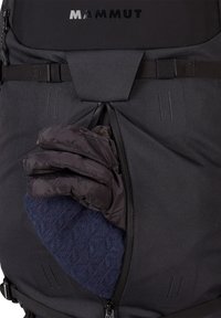 Czarny plecak Mammut z zamykaną kieszenią, w której znajduje się ręka trzymająca ciemnoniebieską teksturowaną rękawicę, wykonaną z izolacyjnego materiału.