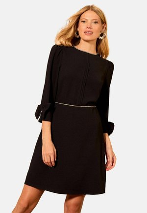 Black dress with long sleeves, pleated detailing, bow accents at the cuffs, and a decorative belt featuring small rhinestones. Smooth fabric texture.