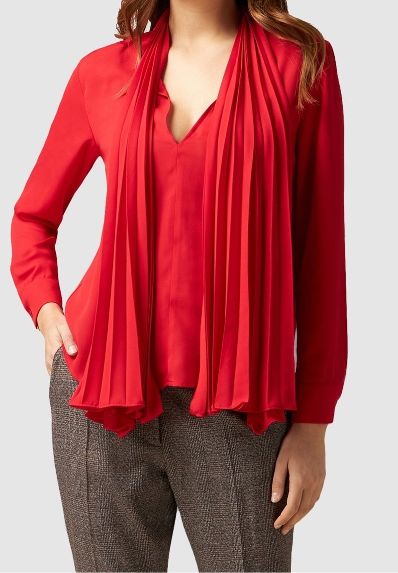 Blouse rouge à manches longues avec un foulard plissé au niveau de l'encolure, confectionnée en tissu léger, associée à un pantalon gris à carreaux texturé.