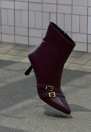 Bottine en cuir bordeaux foncé à talon haut, bout pointu et deux brides à boucle dorée sur une surface pavée près d’un mur carrelé.