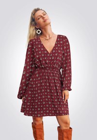 Bordeauxfarbener Blumenmaxikleid mit V-Ausschnitt, langenÄrmeln und elastischem Bund. Hat einen gerafften Rock und wird mit kniebewährten braunen Stiefeln kombiniert.