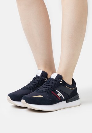 Zapatillas azul marino con parte superior de ante y textil, suela de goma blanca, acentos dorados y rayas rojas y blancas en el costado. Diseño con cordones.