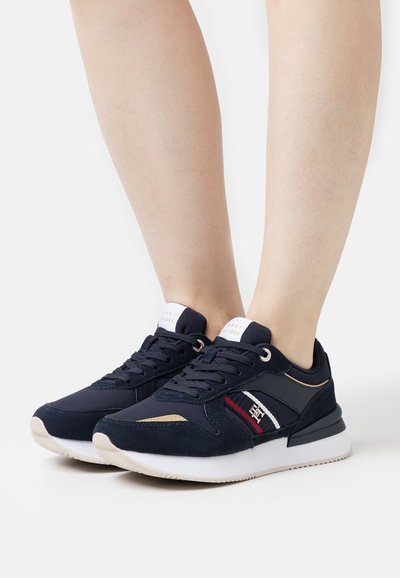 Navy blue sneakers with suede and textile upper, white rubber sole, gold accents, and red and white stripes on the side. Laced design.