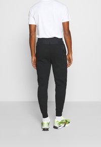 Černé teplákové kalhoty se zúženým střihem, elastickým pasem a boční kapsou na zip, v kombinaci s výraznými zeleno-černými teniskami.