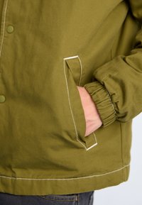 Veste vert olive en tissu texturé ; elle dispose d'une fermeture à pression, de poignets élastiques et d'une poche latérale avec des détails de couture blanche.