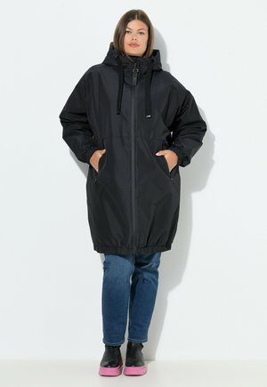 Black long raincoat with a hood, elastic cuffs, and a cinched waist. Features side pockets and a zip front closure. Worn with blue jeans and black shoes.