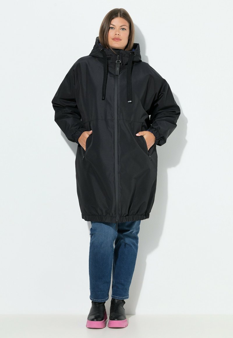 Zwarte lange regenjas met een capuchon, elastische boorden en een getailleerde taille. Voorzien van zijzakken en een ritssluiting aan de voorkant. Draag je met blauwe jeans en zwarte schoenen.