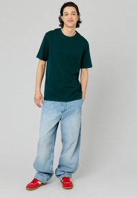 T-shirt en coton vert foncé à manches courtes, associé à un jean en denim baggy bleu clair et des baskets rouges. Design simple sans motifs visibles.