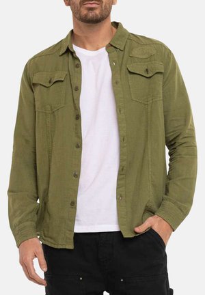 Mand iført olivengrønt skjortebluse over hvid t-shirt og sorte bukser, hånd i lommen, mod en ensartet lys baggrund.