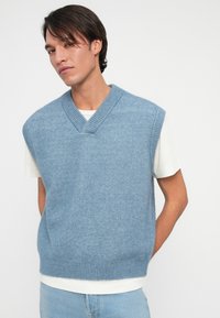 Jovem com cabelo escuro, vestindo um colete de malha azul claro sobre uma t-shirt branca e jeans azul claro, em pé com as mãos atrás das costas.