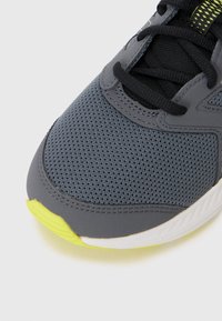 Chaussure de sport grise avec une tige en mesh respirant, des lacets noirs et un accent jaune néon sur la semelle extérieure. Texture lisse avec un col rembourré.