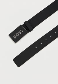 Zwarte synthetische riem met een rechthoekige metalen gesp waarop het woord "BOSS" staat in gepolijste letters. De riem heeft meerdere verstelgaten.