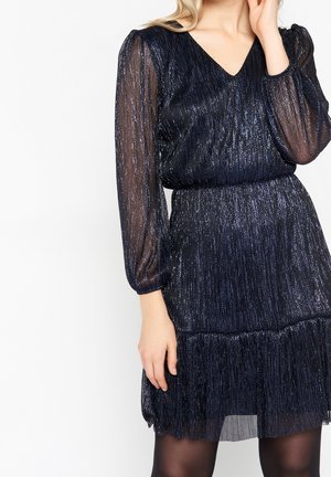 Kobieta ubrana w granatową, błyszczącą suknię z przezroczystymi długimi rękawami i marszczonym stanem, stojąca na jednolitym białym tle.