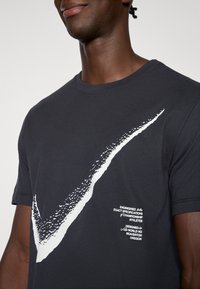 Μαύρο μπλουζάκι με μεγάλο λευκό γραφικό σχήμα (swoosh) και κείμενο στην αριστερή πλευρά. Κοντά μανίκια, στρογγυλή λαιμόκοψη και απαλό ύφασμα.