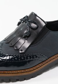 Gros plan sur une chaussure brogue en cuir noir avec perforations décoratives, détail à franges, nœud sur le dessus et semelle en caoutchouc brun à crampons.
