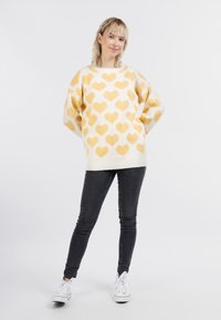 Maglione lavorato a maglia con base crema e motivi a cuore gialli, design over-sized, scollatura ampia e polsini a coste, abbinato a jeans neri e sneakers bianche.