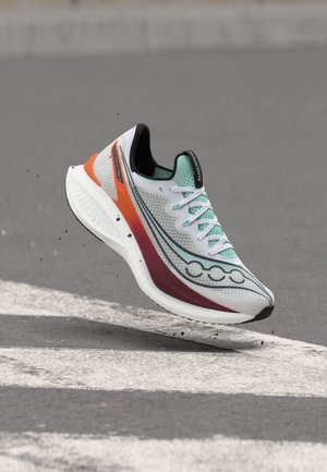 Loopschoen met witte, petrolblauwe, oranje en mahonie kleuren zwevend boven een grijs asfaltoppervlak met witte wegmarkeringen.