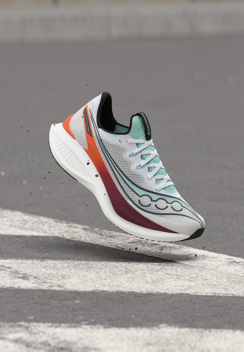 Αθλητικό παπούτσι με λευκό, τιρκουάζ, πορτοκαλί και μαρόν χρώμα να αιωρείται πάνω από μια γκρίζα ασφάλτινη επιφάνεια με λευκές οδικές σημάνσεις.