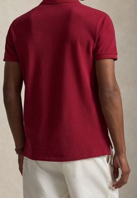 Homme portant un polo rouge foncé à manches courtes et un pantalon blanc, vu de dos sur un fond neutre uni.