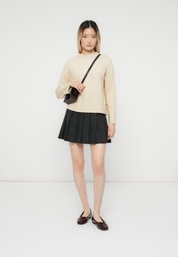 Pull en maille beige, jupe plissée noire et ballerines bordeaux. Tenant un petit sac à bandoulière noir. Tenue simple et moderne sur un fond uni.