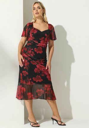 Vestido negro con flores rojas, mangas cortas con volante, cuerpo fruncido, dobladillo transparente y silueta ajustada. Llevado con tacones negros con tiras.